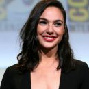 Actress Gal Gadot wears a dark top and smiles wearing red lipstick.