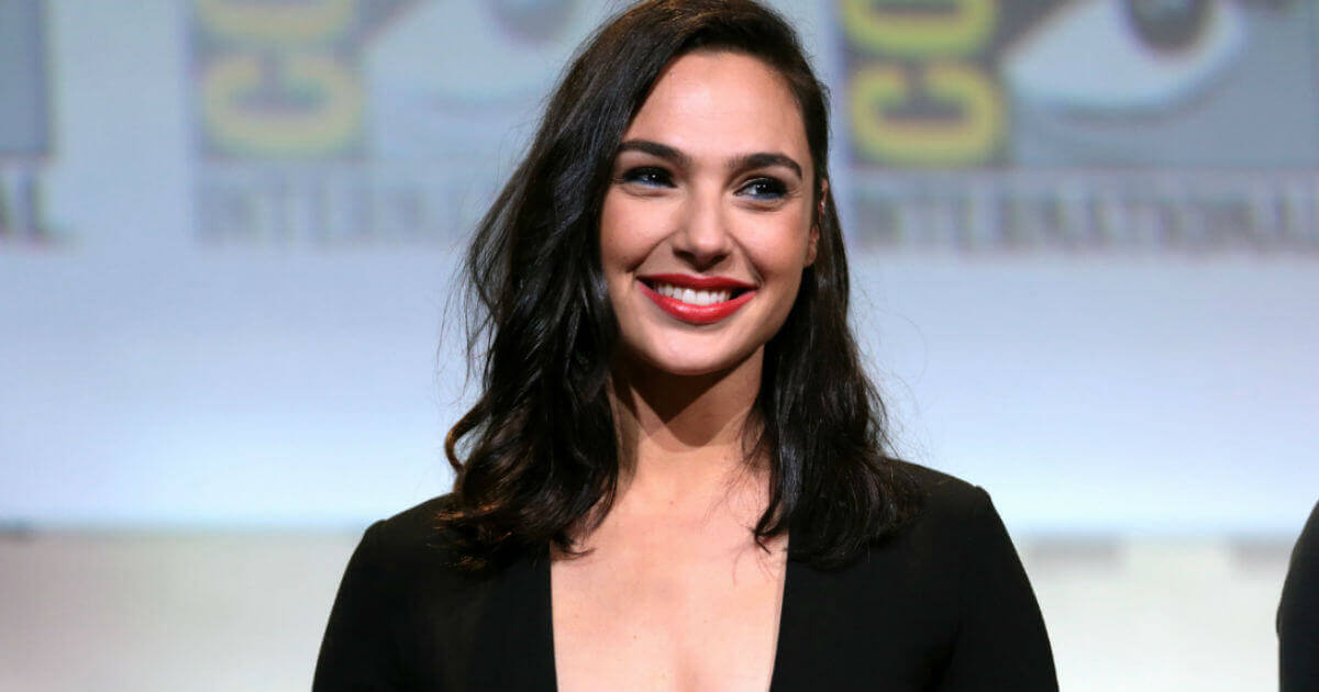 Actress Gal Gadot wears a dark top and smiles wearing red lipstick.