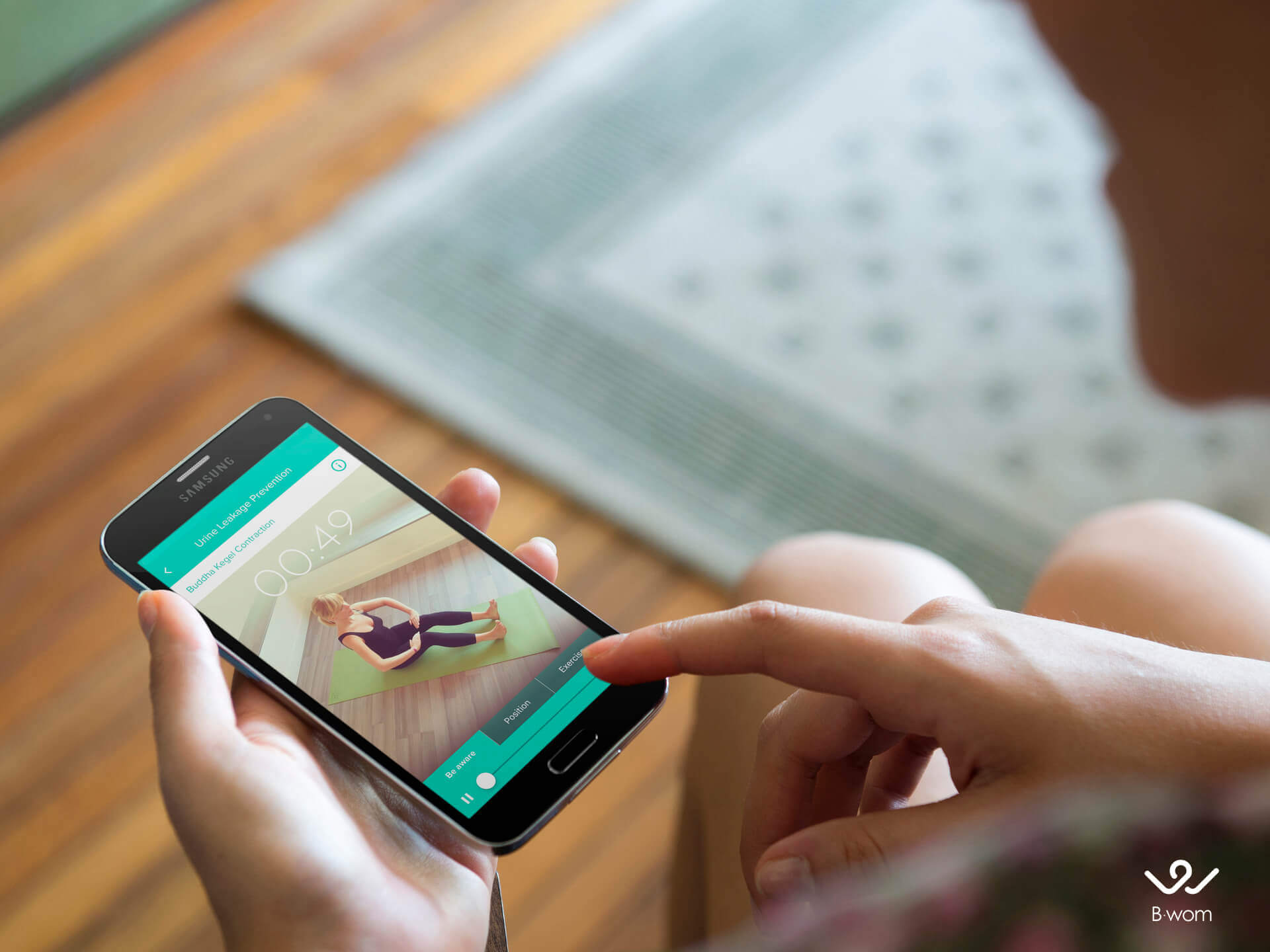 A woman's hands hold a smartphone that has the B-wom app open showing a woman doing excercise.