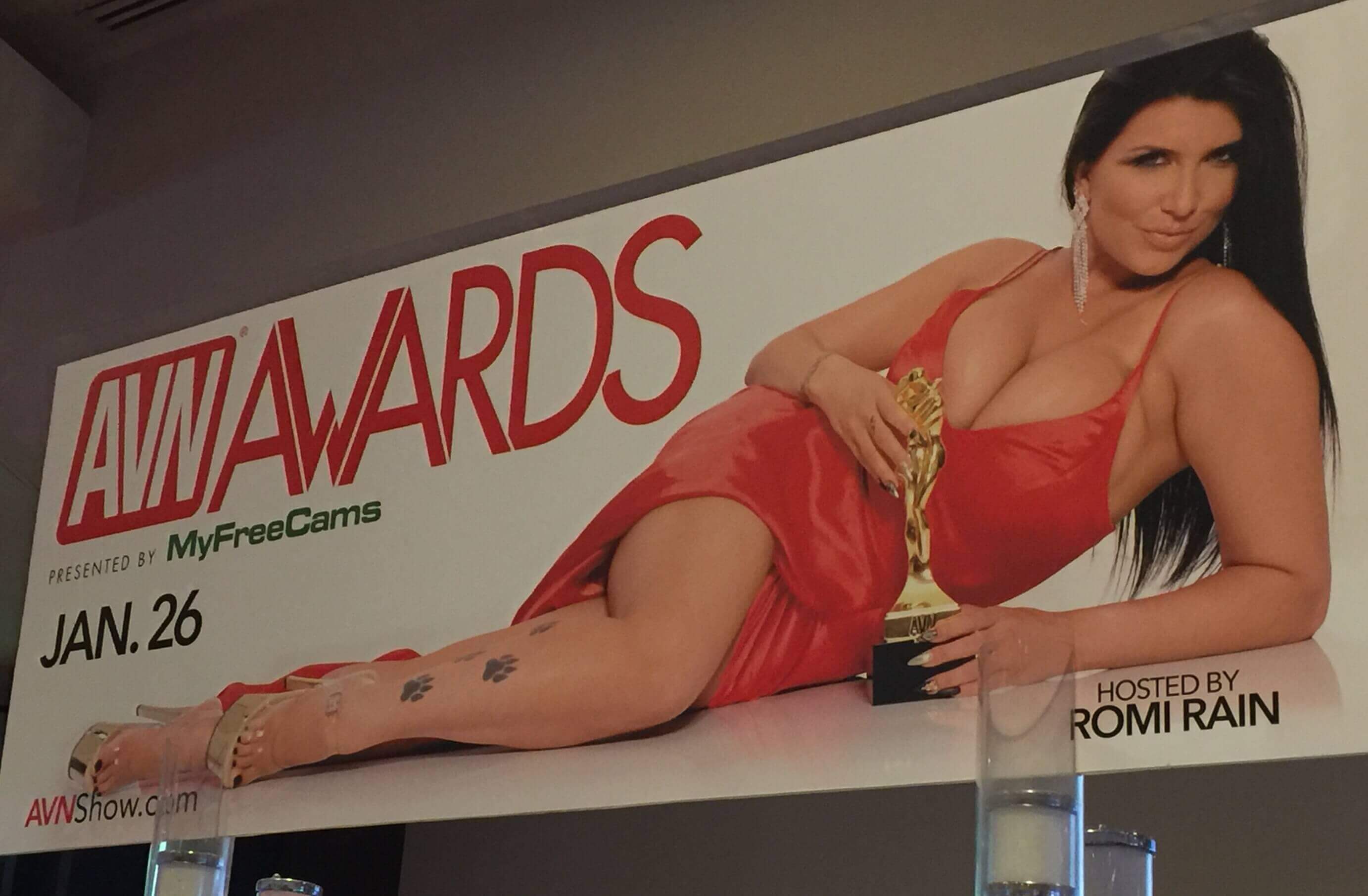 A brunette woman wearing a revealing orange dress lies on her side with an gold awards trophy next to the text AVN Awards 2019.