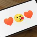 A smartphone shows two heart emojis with a yellow-face kiss emoji in the middle.