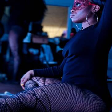 A dancer in fishnet stockings sits in front of a lighted mirror while other coworker getting ready