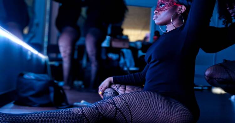 A dancer in fishnet stockings sits in front of a lighted mirror while other coworker getting ready