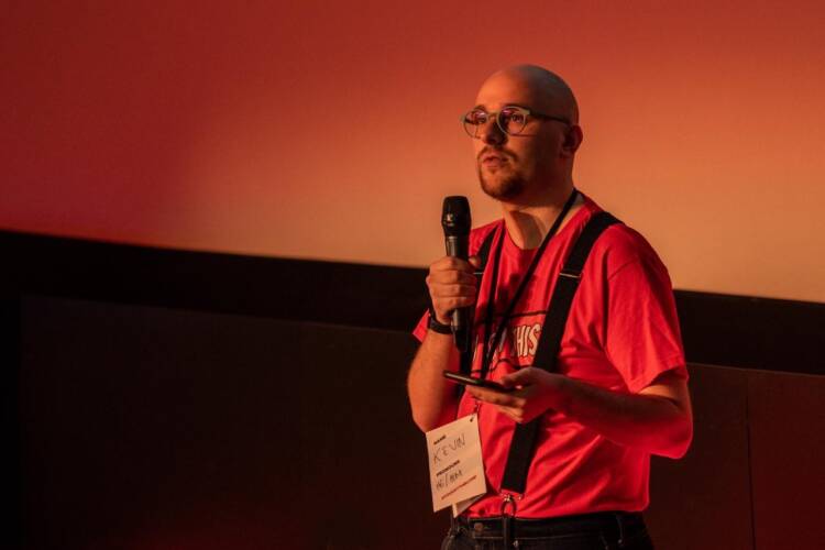 Kevin Lewis introduces the You Got This Birmingham 2020 conference. He is wearing glasses and an orange t-shirt with the You Got This logo across the front. He is looking up into the audience as he holds his phone in his left hand and the microphone in his right.