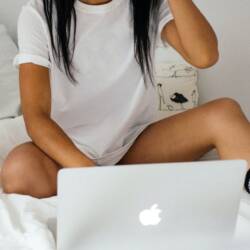 Young woman in white bed wearing black socks watching a Mac computer screen.
