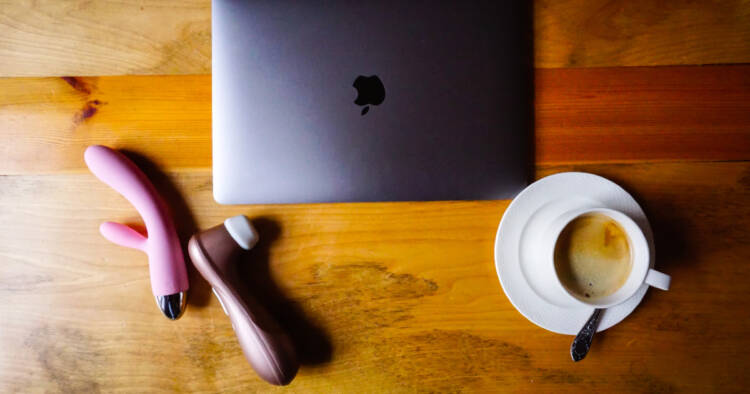 Sex toys sit next to a closed laptop and coffee on a wooden tabletop.