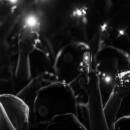 A black and white photo of a crowd at a concert or event. Everyone has their hands raised and most are holding cell phones. The phones have their flashlight setting turned on.