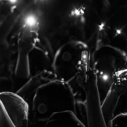 A black and white photo of a crowd at a concert or event. Everyone has their hands raised and most are holding cell phones. The phones have their flashlight setting turned on.