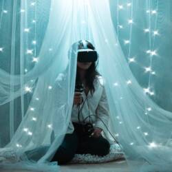 A decorative image of a woman sitting in a canopy wearing a virtual reality headset. She's surrounded by pillows and twinkly lights.