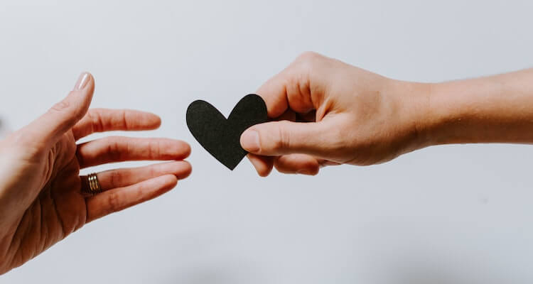 Decorative image of two hands reaching towards each other. On one, you can see a wedding ring. The other is holding a black paper heart.