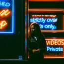 A woman in a leather jacket standing outside a storefront at night. It's dark all around, but you can see the neon signs with sayings like "Licensed Sex Shop", "Strictly over 18s only" and "private videos".