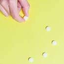Light yellow background with a trail of small round white pills moving up the center of the image. There's a light-skin human hand reaching into frame from the top-left corner grabbing one of the pills.