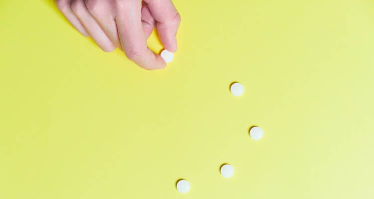 Light yellow background with a trail of small round white pills moving up the center of the image. There's a light-skin human hand reaching into frame from the top-left corner grabbing one of the pills.