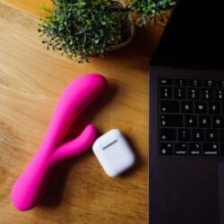 A pink rabbit vibrator and AirPods case laying on a table next to an open laptop.