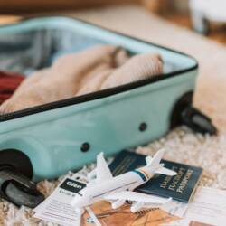 A suitcase open on the floor mid-packing. You can see sweaters inside the suitcase and a passport and model airplane next to it.