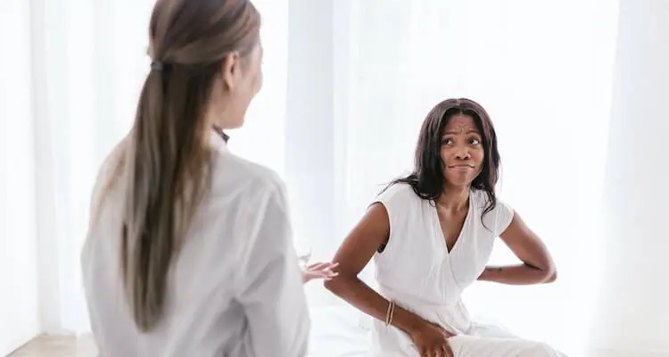A doctor talking to a sitting patient. The patient has an uncomfortable face and holding their lower back.