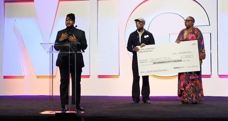 Glenise giving an acceptance speech with Pharrell Williams and CEO Felecia Hatcher holding the check in the background.
